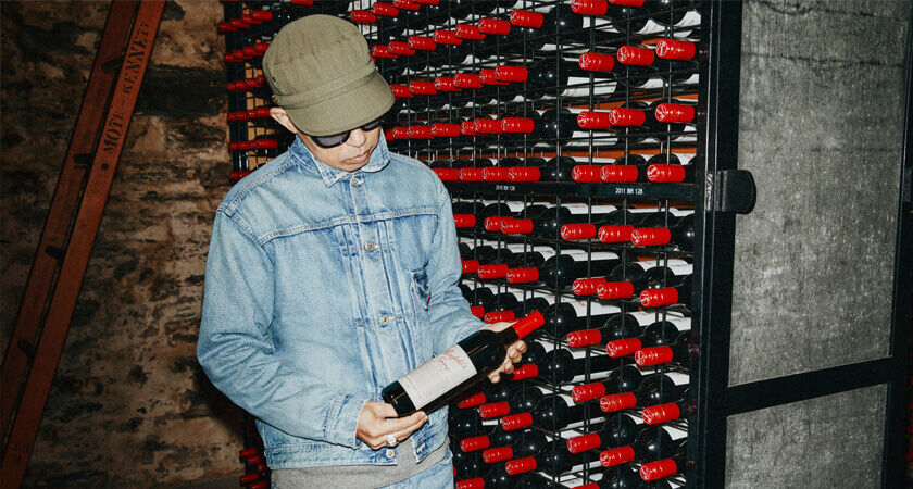 Nigo holds bottle of Grange in cellar with red bottles on shelf behind.
