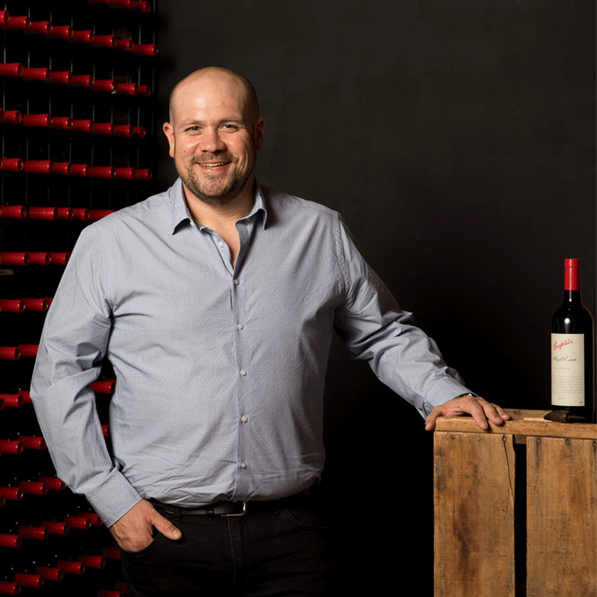 Matt Woo, Penfolds Winemaker, stands with a bottle of wine beside him and rows of red Penfolds bottles behind